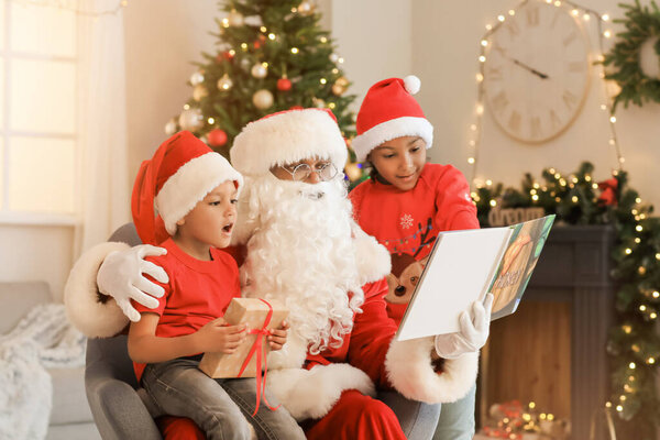 African-American Santa Claus with cute children reading book at home on Christmas eve