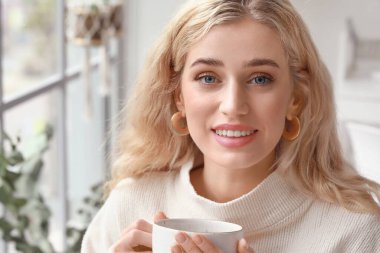 Güzel, genç bir kadın sıcak kıyafetler içinde evde çay içiyor.