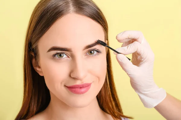 Young woman undergoing eyebrow correction procedure on color background