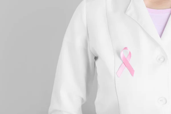 Female doctor with pink ribbon on grey background, closeup. Breast cancer awareness concept