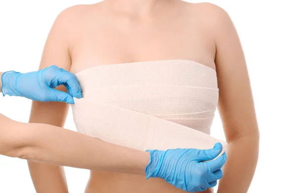 Doctor applying bandage on female chest after cosmetic surgery operation against white background