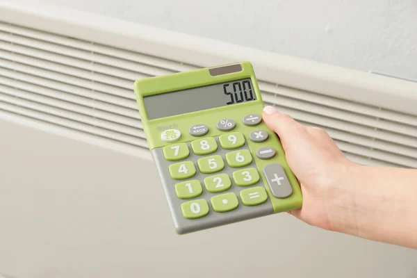 Woman with calculator near radiator, closeup. Concept of heating season