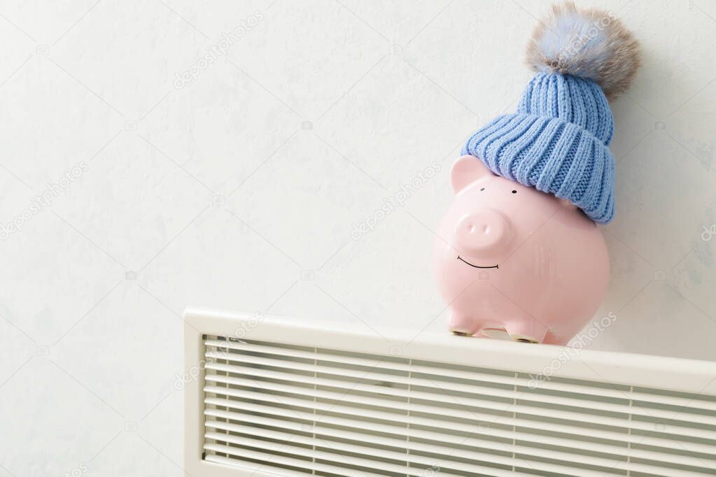 Piggy bank and hat on radiator. Concept of heating season