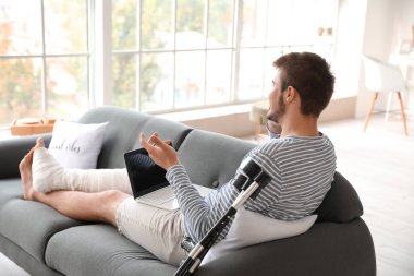 Kırık bacaklı genç adam evde telefonla konuşurken dizüstü bilgisayar kullanıyor.