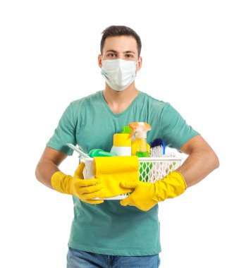 Tıbbi maskeli genç bir adam ve beyaz arka planda temizlik malzemeleri var.