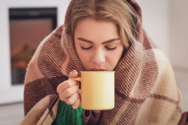 Sıcak ekoseye sarılı genç bir kadın evde sıcak çay içiyor. Isınma mevsimi kavramı
