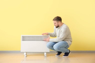 Evdeki elektrikli ısıtıcının yanındaki genç adam. Isınma mevsimi kavramı