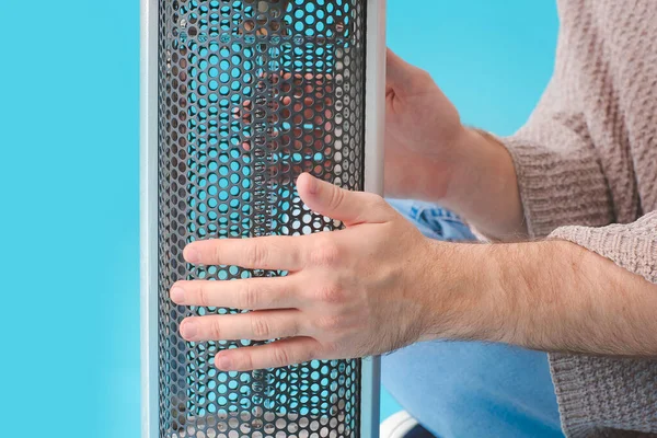 Young man warming hands near electric heater on color background, closeup. Concept of heating season