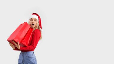 Noel Baba şapkalı, açık arka planda alışveriş torbalarıyla şaşırmış genç bir kadın.