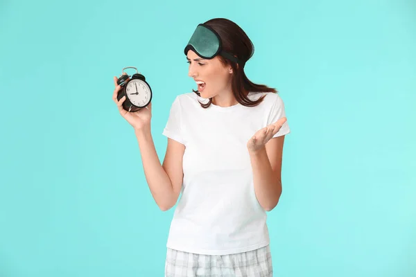 Emotional woman with sleep mask and alarm clock on color background