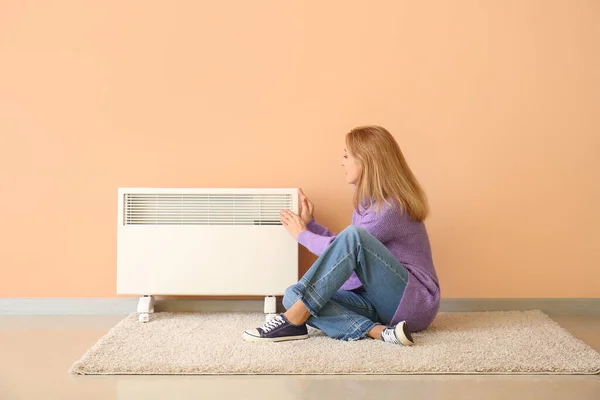 Mature woman near electric heater at home. Concept of heating season