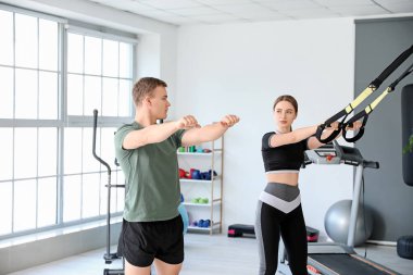 Koç, spor salonunda genç bir kadının TRX kayışlarıyla antrenman yapmasına yardım ediyor.