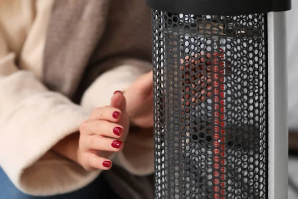 Young woman warming hands near electric heater at home. Concept of heating season