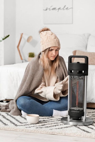 Young woman in warm clothes sitting near electric heater at home. Concept of heating season