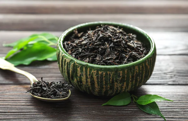 Dry black tea leaves in bowl and spoon on wooden background