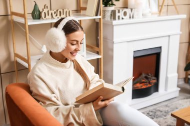 Evde şöminenin yanında kitap okuyan genç bir kadın.