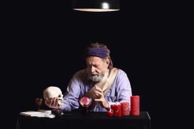 Male fortune teller with crystal ball and human skull at table on dark background clipart