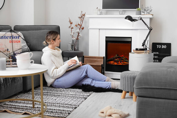 Young woman reading book near fireplace at home