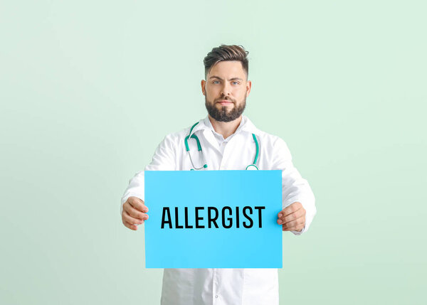 Portrait of male doctor holding poster with text ALLERGIST on color background