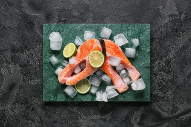 Koyu arkaplanda çiğ somon bifteği, limon ve buz.