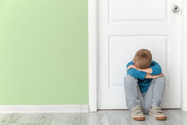 Üzgün küçük çocuk kapalı kapının yanında oturuyor.