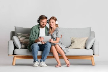 Tablet bilgisayarı ve kulaklıkları olan genç bir çift, arka planda ışığa karşı kanepede oturuyor.