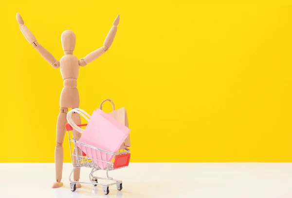 色の背景にショッピングカートやバッグと木製のマネキン — ストック写真