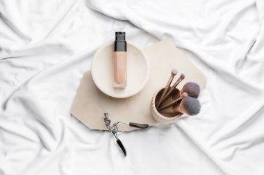 Kumaş arka planında bir şişe makyaj vakfı, fırçalar ve kirpik bigudisi.