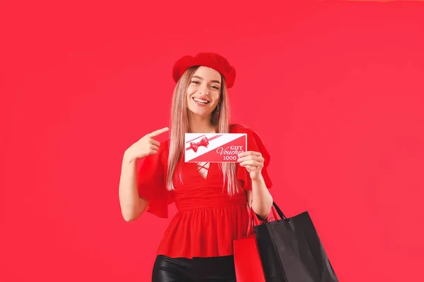 Beautiful young woman with gift voucher and shopping bags on color background