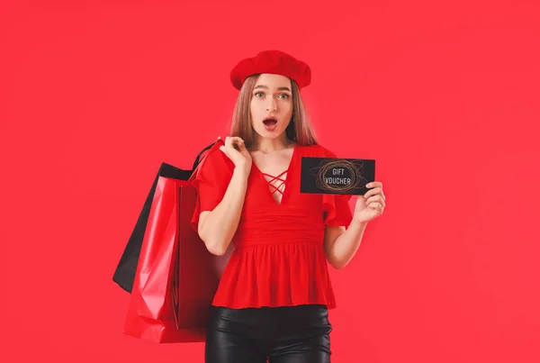 Surprised young woman with gift voucher and shopping bags on color background