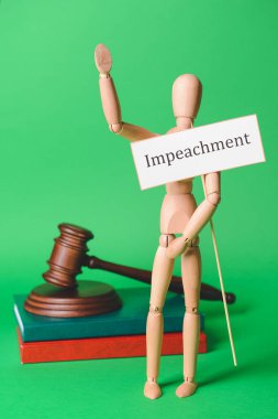 Tahta manken elinde IMPEACHMENT sözcüğü, kitaplar ve renk arkaplanı hakkında hakim tokmağı olan bir kağıt tutuyor.