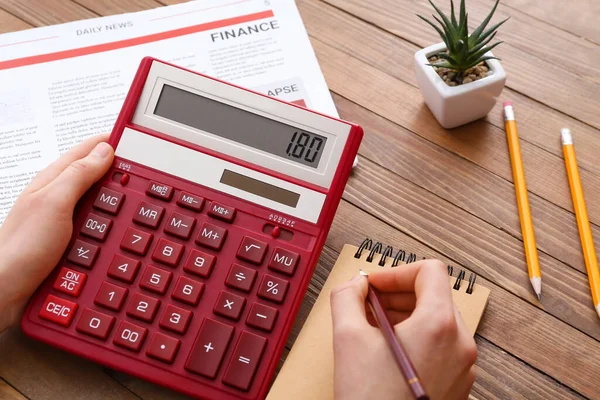 Woman with calculator writing in notebook at wooden table