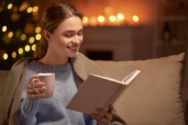 Güzel genç bir kadın sıcak çikolata içiyor ve Noel arifesinde evde kitap okuyor.