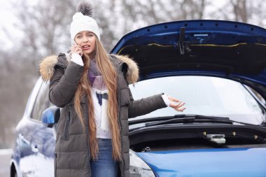 Stresli genç bir kadın dışarıda kırık bir arabanın yanında telefonla konuşuyor.