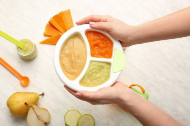Woman holding plate with different baby food on light background