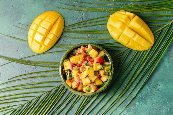 Bowl with fresh mango salsa and tropical leaf on color background