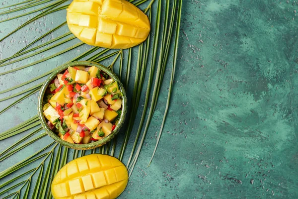 Bowl with fresh mango salsa and tropical leaf on color background