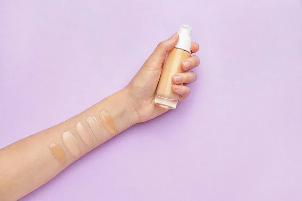 Female hand with bottle of makeup foundation and samples on color background