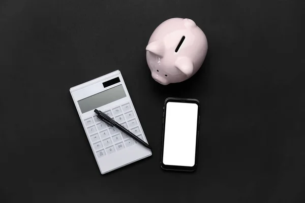 Calculator with mobile phone and piggy bank on dark background