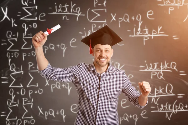 Happy male student in graduation hat and with diploma near blackboard