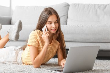 Evde dizüstü bilgisayarı olan genç bir kadın cep telefonuyla konuşuyor.