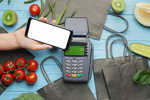 Female hand with mobile phone, payment terminal and food on color wooden background