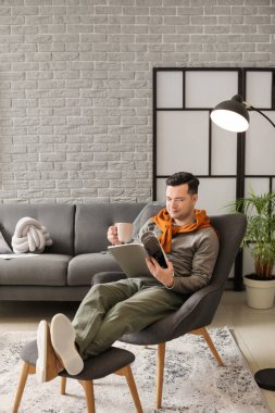 Genç adam evde koltukta otururken dergi okuyor ve çay içiyor.