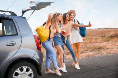 Mutlu genç kadınlar arabanın yanında selfie çekiyorlar.