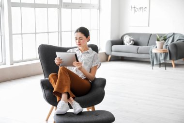 Tablet bilgisayarlı güzel genç bir kadın evdeki koltukta oturuyor.