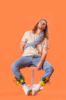 Çiçekli yakışıklı bir gencin yaratıcı portresi renk arka planına karşı taburede oturuyor.