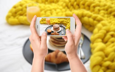 Cep telefonlu kadın yemek fotoğrafçısı yatakta lezzetli bir kahvaltı fotoğrafı çekiyor.