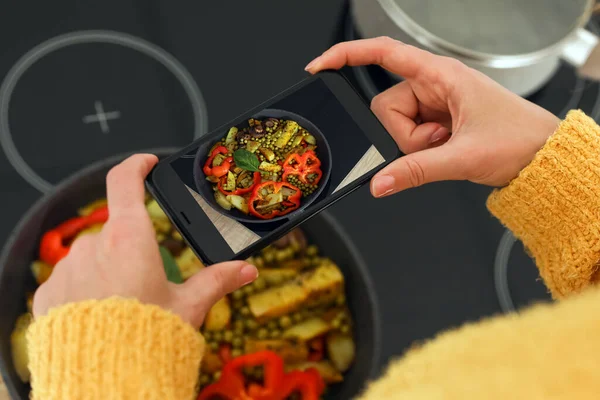 Female food photographer with mobile phone taking picture of tasty fried vegetables in pan on stove, top view