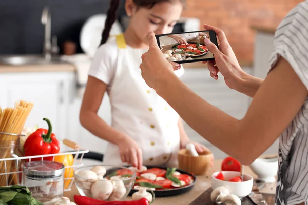 Female food photographer with mobile phone and little girl cooking pizza in kitchen