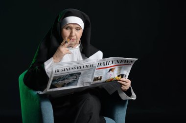 Gazetesi ve sigarası olan yaşlı rahibe karanlık arka planda koltukta oturuyor.